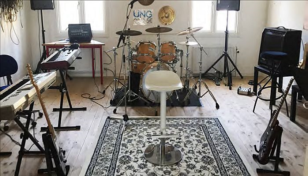 Book et øvelokale MusikLaboratoriet fls. Billede af øvelokale med keyboards, bas, guitar, trommesæt, højtalere og en stol, der står på et tæppe i midten af lokalet.