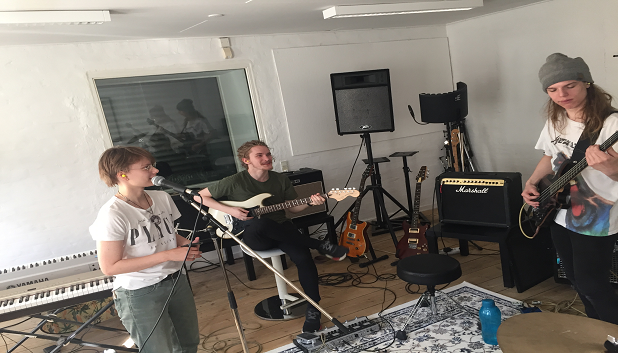 Band. Billedet viser tre unge i et øvelokale, én står op og spiller bas, én står og synger og én sidder og spiller guitar.