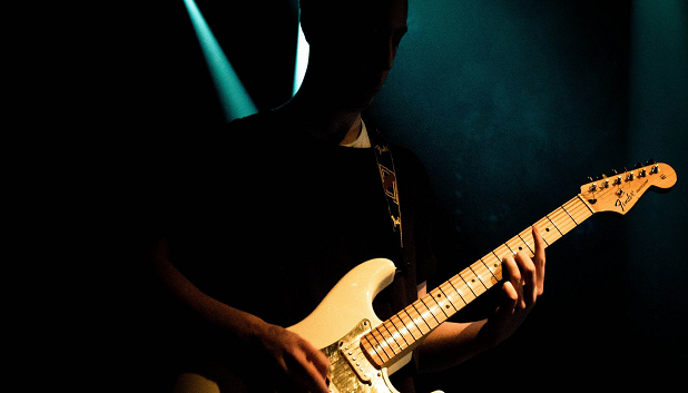 Guitar. Billedet viser en person, der står i skygge, og spiller på en guitar, hvor der er spot på.